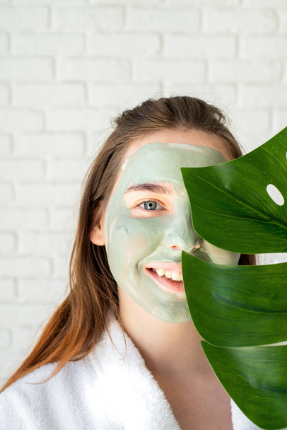 スパとウェルネス。天然化粧品。セルフケア。モンスターの葉を持つ顔のマスクを持つ幸せな若い女性の肖像 - 写真・画像