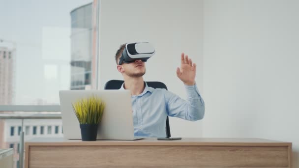 Un joven sentado en un escritorio de la oficina utiliza gafas de realidad aumentada para trabajar en proyectos empresariales en varios campos. - Imágenes, Vídeo