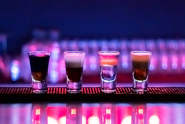 Bebidas cortas en el bar nocturno. Cuatro vasos de cócteles alcohólicos. Luz de neón colorida, sombras oscuras. - Foto, Imagen