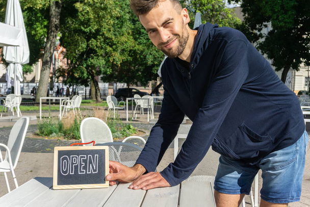 Waiter with Open sign on table open-air cafe. Smiling barista holding sign Open. Cafe owner welcome customers. Reopen for business. - Photo, Image