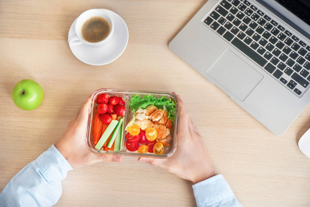 Gezond zakenlunchen op kantoor. Zakenman met groente- en kipsalade in lunchbox op houten bureau vlakbij laptop. Bovenaanzicht. - Foto, afbeelding