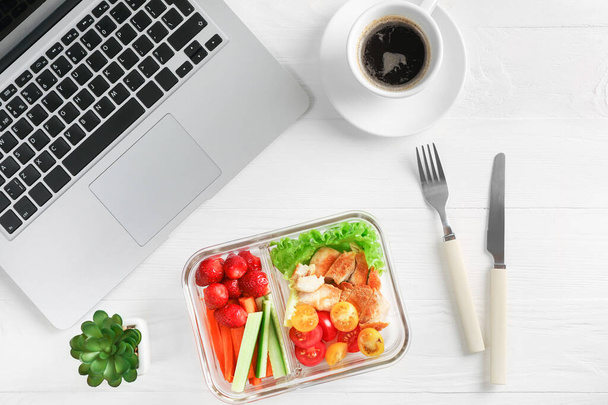 Zdravý pracovní oběd v kanceláři. Horní pohled na zeleninový a kuřecí salát v krabici na oběd a šálek kávy na bílém dřevěném stole poblíž notebooku. - Fotografie, Obrázek