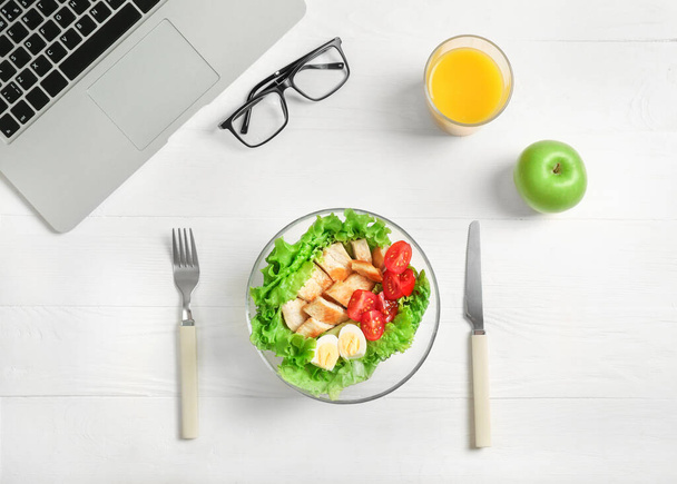 Healthy business lunch in office workplace. Top view of vegetable and chicken salad in bowl, fresh green apple, glass of orange juice on white wooden desk near laptop and glasses. - Foto, immagini