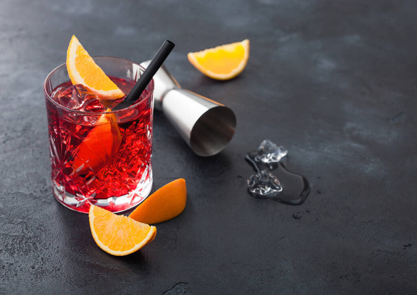 Negroni cocktail in crystal glass with orange slice and fresh raw oranges with jigger and ice cubes on black background. Top view - Фото, изображение