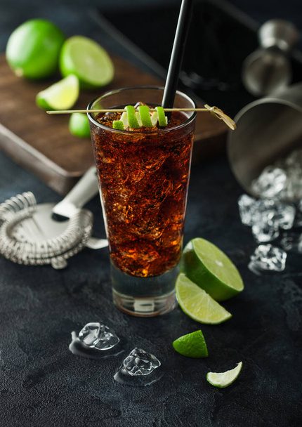 Cuba Libre cocktail in highball glass with ice and lime peel on bamboo stick with straw and fresh limes on black table background. Shaker and strainer on bar stand. Top view - Fotografie, Obrázek