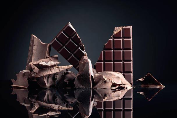 A broken chocolate bar and pieces of dark chocolate on a black reflective background. Copy space. - Foto, Imagem