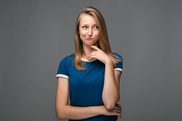 Mulher jovem pensativo com cabelo liso loiro em uma camisa azul está de pé com a mão levantada no queixo e olhando atenciosamente longe, isolado sobre fundo cinza. Conceito de pensamento - Foto, Imagem