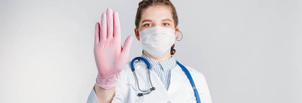A young beautiful woman doctor in a white coat and pink sterile gloves shows a stop gesture on white background. Say No virus disease. - Photo, Image