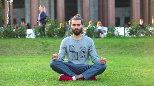 Vousáč medituje o zelené trávě v městském parku. Koncept. Stylový mladý muž medituje o zelené trávě v rušném městě. Krásný stylový muž se zajímá o meditaci - Záběry, video