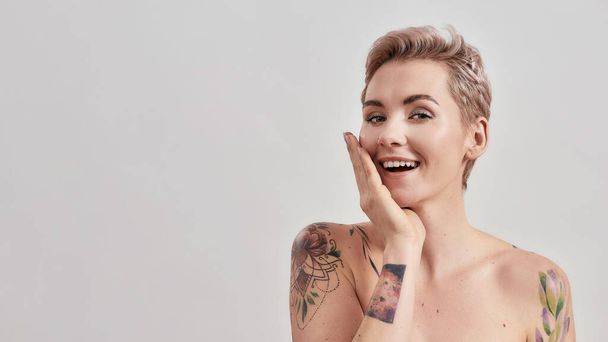Portrait of half naked tattooed woman with short hair smiling at camera, touching her face with one hand isolated over light background - Фото, изображение