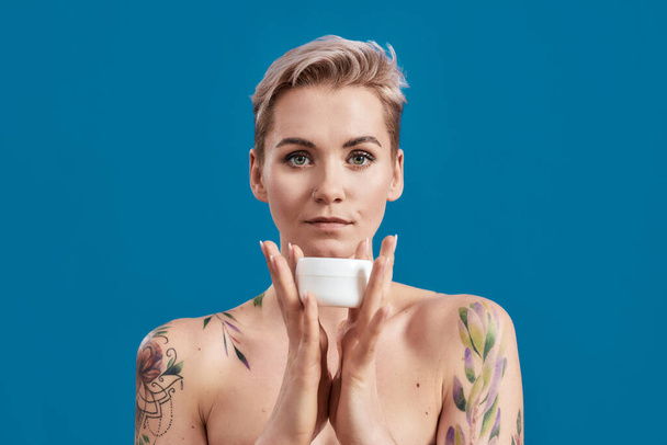 Portrait of beautiful half naked tattooed woman with short hair holding white plastic jar of cream or body lotion isolated over blue background - Foto, afbeelding