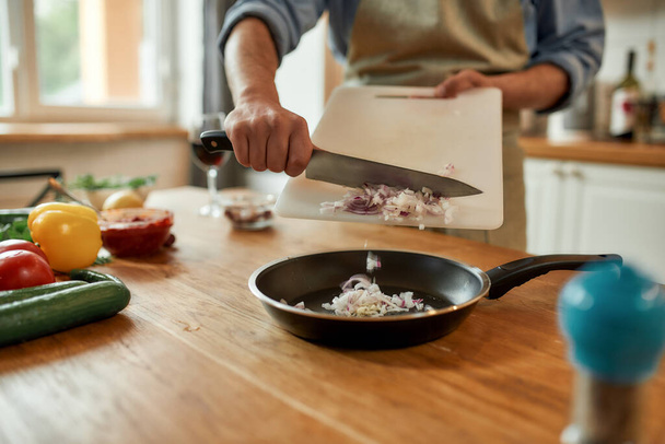 Close up of man in apron holding chopping board, putting onion in the pan for frying while cooking dinner. Healthy nutrition, cooking at home concept - Foto, imagen