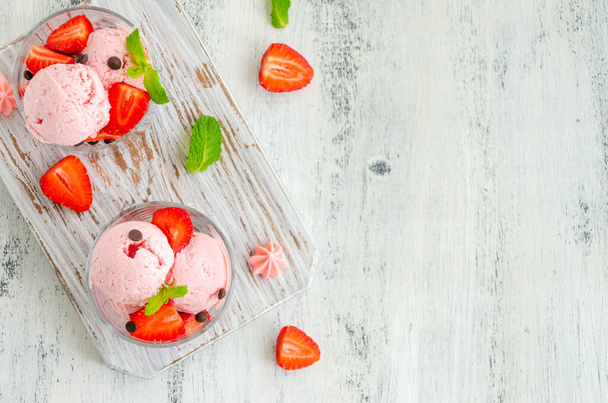 Strawberry ice cream in a glass with fresh strawberries, chocolate drops and mint on a board on a light wooden background. Horizontal, copy space, top view - Photo, Image
