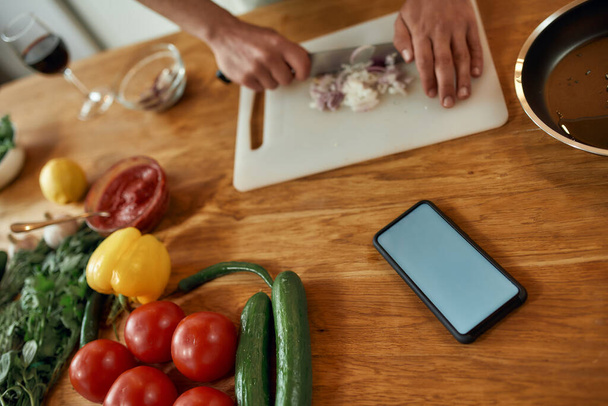 Close up of phone with blank screen on the table. Man cutting onion on chopping board, using smartphone app while cooking dinner. Healthy nutrition, technology concept - Foto, afbeelding