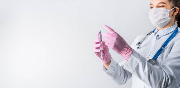Een jonge vrouwelijke arts in een witte jas, met een medisch masker en roze steriele handschoenen houdt de spuit vast. Focus op de spuit. Inentingsconcept. - Foto, afbeelding