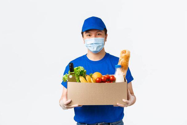 Online αγορές, διανομή τροφίμων και covid-19 πανδημία έννοια. Φιλικός χαμογελαστός ασιάτης κούριερ με ιατρική μάσκα, γάντια και μπλε στολή φέρνει παραγγελία τροφίμων στον πελάτη, παραδίδοντας κουτί στον πελάτη - Φωτογραφία, εικόνα