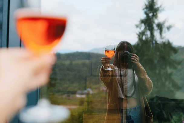 Frau hält Aperolgetränk in der Hand und macht Selfie im großen Fenster vor dem Hintergrund von Bergen, Sommerurlaub und Ferienort. Frau jubelt mit leckerem Orangencocktail - Foto, Bild