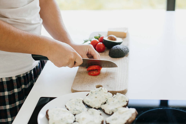 Osoba krojąca plasterek pomidora na kanapkę w nowoczesnej białej kuchni. Proces tworzenia zdrowych toastów z awokado, pomidory, rukola, ser i chleb pełnoziarnisty. Koncepcja gotowania domowego - Zdjęcie, obraz