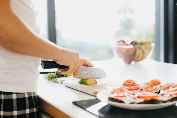 Osoba krájí avokádo na sendvič v moderní bílé kuchyni. Proces výroby zdravých toastů s avokádem, rajčaty, rukolou, sýrem a celozrnným chlebem. Domácí kuchyně koncept - Fotografie, Obrázek