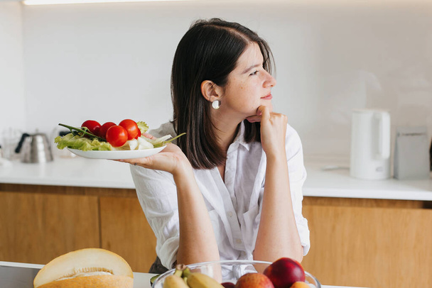 Terveellistä ruokaa. Nuori onnellinen nainen tilalla levy vihreä salaatti, rucola ja kirsikkatomaatti taustalla moderni valkoinen keittiö. Kotiruokaa, salaatin ainesosia - Valokuva, kuva