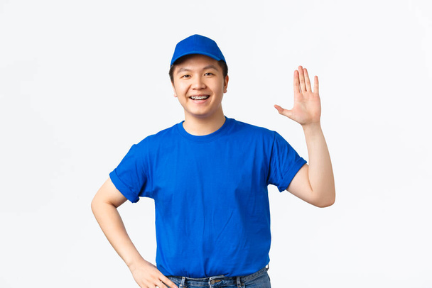 Mavi üniformalı arkadaş canlısı Asyalı erkek kurye gülümseyip elini sallayarak merhaba diyor. Selamlayan müşteri müşteriyi kapısına getiriyor. Neşeli teslimatçı, selam jest, beyaz arka plan - Fotoğraf, Görsel