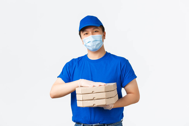 Contactless delivery, covid-19 shipping, preventing virus concept. Smiling friendly asian male courier in medical mask, gloves and blue uniform bring pizza order to client house, white background - Photo, Image