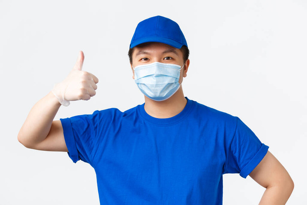 Online shopping, delivery and coronavirus pandemic, preventing virus concept. Close-up of upbeat smiling asian male courier in medical mask, gloves and blue uniform, showing thumbs-up - Photo, Image