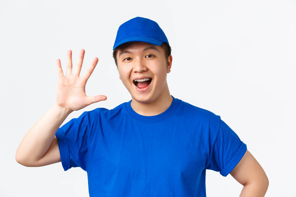 Online nakupování, rychlé dodání, zaměstnanci a domácí dodávky koncept. sebevědomý usměvavý asijský kurýr s rovnátky, v modré uniformě, ukazující číslo pět, bílé pozadí - Fotografie, Obrázek