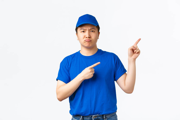 Αναστατωμένος και μελαγχολικός Ασιάτης κούριερ με μπλε καπέλο και t-shirt να γκρινιάζουν απογοητευμένοι και να δείχνουν στην πάνω δεξιά γωνία. Λυπηρός ντελιβεράς με στολή ενημερώνει άσχημα νέα, στέκεται λευκό φόντο - Φωτογραφία, εικόνα