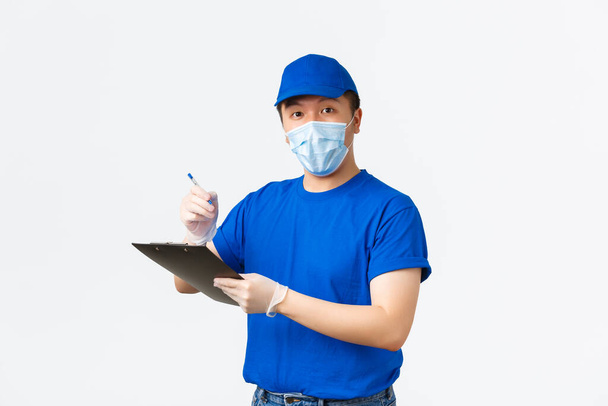 Contactless delivery, covid-19 shipping, preventing virus concept. Friendly-looking asian male courier in blue uniform, medical mask and gloves look up clients address during shipment, hold clipboard - Photo, Image