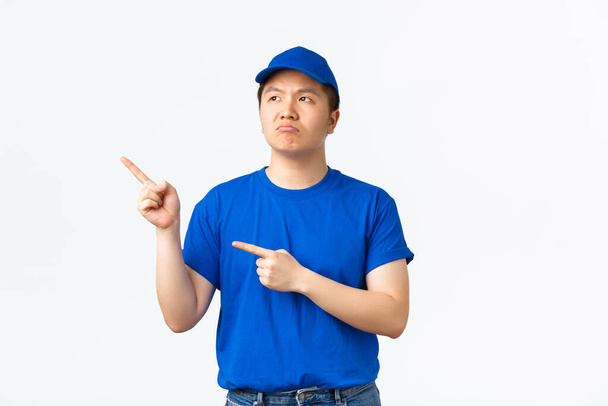 Escéptico y decepcionado mensajero asiático en uniforme azul quejándose de algo malo. Repartidor en gorra y camiseta apuntando esquina superior izquierda y mirando con arrepentimiento, fondo blanco - Foto, imagen