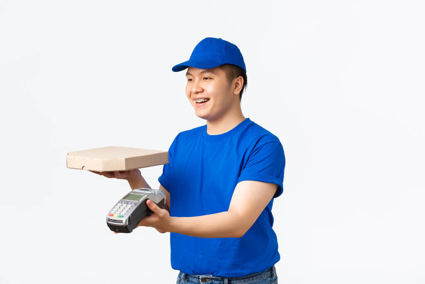 Kontaktloses Einkaufs-, Bezahl- und Lieferkonzept. Freundlich lächelnder asiatischer Kurier in blauer Uniform, Pizzaschachtel aushändigen, Bestellung an den Kunden geben und Kassenterminal, weißer Hintergrund - Foto, Bild