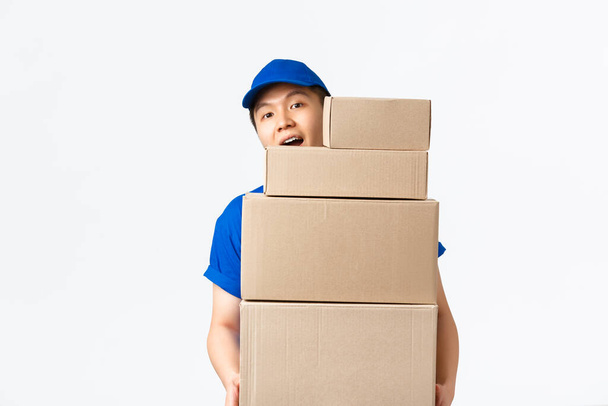Интернет-шопинг, концепция быстрой доставки. Молодой азиатский курьер в синей форме несет коробки с заказами клиентов, приносит доставку в дом клиента, глядя на камеру веселый, белый фон - Фото, изображение