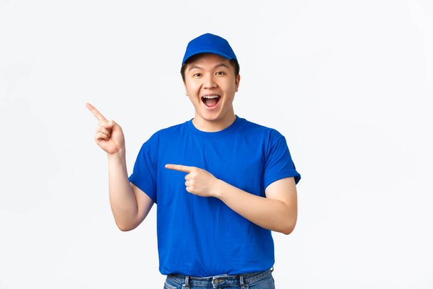 Veselý mladý asijský kurýr v modré uniformě ukazující prsty v levém horním rohu, reklama na přepravní společnost, zásilka balíků. Roztomilý doručovatel něco propagovat - Fotografie, Obrázek