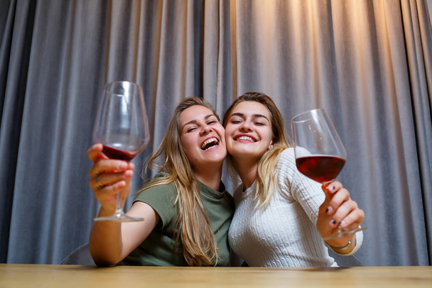 zwei Mädchen europäischen Aussehens mit blonden Haaren sitzen am Tisch, trinken Wein und lachen, entspannen zu Hause, Alkohol - Foto, Bild