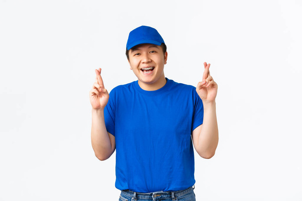 Retrato de emocionado chico asiático esperanzado con aparatos ortopédicos, repartidor en azul uniforme esperanza y pedir deseos, dedos cruzados buena suerte. Mensajero mirando feliz y sonriente mientras reza, esperando resultados - Foto, Imagen