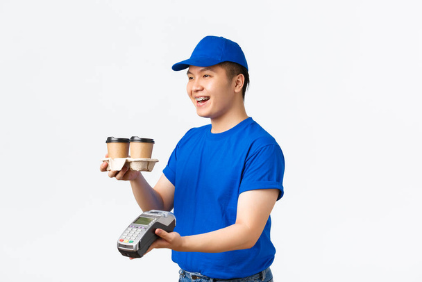 Contactless shopping, payment and delivery concept. Side view of pleasant smiling asian courier in blue uniform, handing takeaway coffee order and POS terminal for paying, white background - Photo, Image