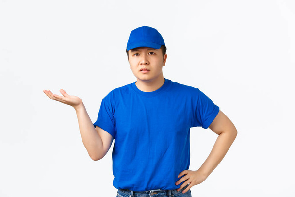 Confundido y molesto repartidor asiático en uniforme azul señalando la mano izquierda, mirando decepcionado, esperando explicación. Mensajero no puede entender algo, de pie fondo blanco - Foto, imagen