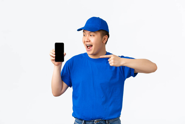 Tecnologia, compras on-line e conceito de transporte. alegre sorrindo asiático courier em azul uniforme, apontando dedo para a tela do telefone móvel, mostrando aplicação, recomendo cliente para rastrear a ordem com app
 - Foto, Imagem