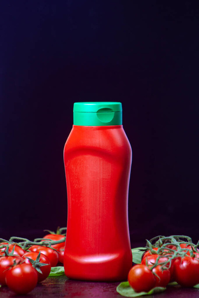 fles, tube rode ketchup met groene dop en twee rijpe takken kerstomaten met spinazie op een zwarte reliëftafel. close up, verticale oriëntatie mockup - Foto, afbeelding