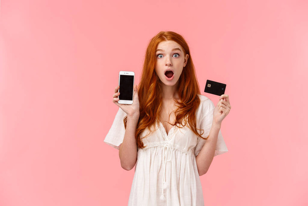 Любопытная и впечатленная рыжая европейская женщина рассказывает об удивительной рекламе, специальных скидках в интернет-магазине, захватить ее кредитную карту, показывая экран смартфона, открытый рот поражен
 - Фото, изображение