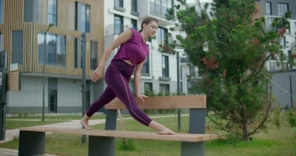 Atletyczna kobieta siedzi na sznurku na ławce w podwórku budynku mieszkalnego, fitness w środowisku miejskim, zajęcia jogi na świeżym powietrzu, ćwiczenia fizyczne w mieście, 4k 120p Prores HQ - Materiał filmowy, wideo