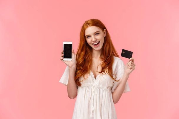 Excited, sassy, carefree female with red curly hair like wasting money in internet store, making order, got tickets in flight app, holding credit card and smartphone, showing promo on mobile screen - Photo, Image