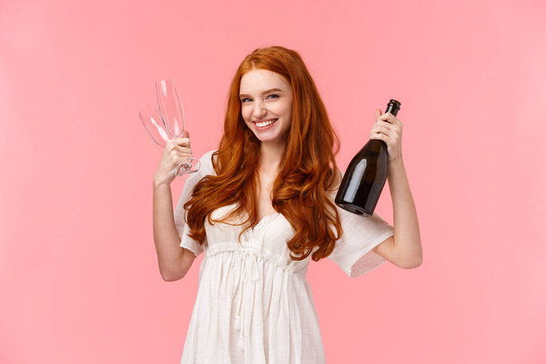Τολμηρή κοκκινομάλλα που γιορτάζει με φιλενάδες, διασκεδάζει μαζί, κρατάει σαμπάνια και δύο ποτήρια, προσκαλεί για ποτό, χαμογελά αναιδής και χαρούμενη, στέκεται ροζ φόντο - Φωτογραφία, εικόνα
