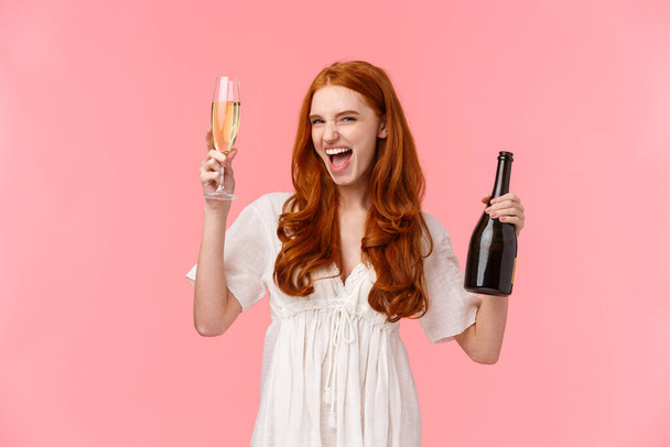 aufgeregt und unbeschwert betrunken rothaarige kaukasische Freundin Spaß haben, wild und frei auf ehrfürchtige Party, heben Glas und Flasche Champagner, frech lächeln, stehen rosa Hintergrund - Foto, Bild