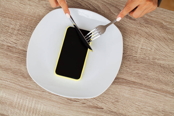 Závislost na telefonu, zavřít ženské ruce drží vidličku a nůž přes telefon na talíři - Fotografie, Obrázek
