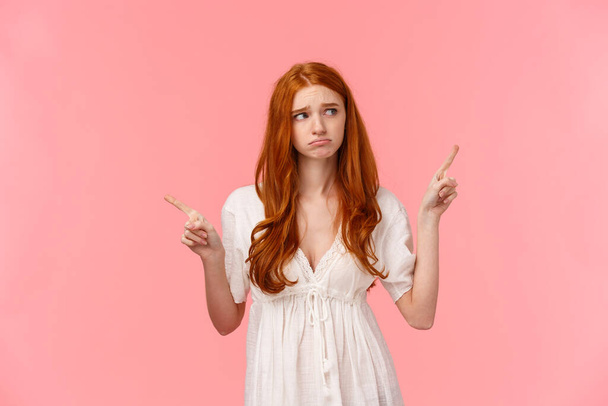 Waist-up portrait innocent, cute and timid redhead indecisive girl, dont know what do, pouting feeling sad, gloomy look around, pointing fingers left and right, pink background - Photo, Image