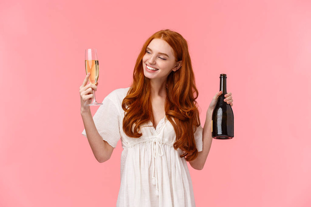 Dziewczyna czuje się zrelaksowana i szczęśliwa lekko pijana uśmiechając się i patrząc na wylewane szkło, trzymając i pijąc szampana, uśmiechając się, imprezując na miesiąc miodowy, stojąc różowe tło - Zdjęcie, obraz