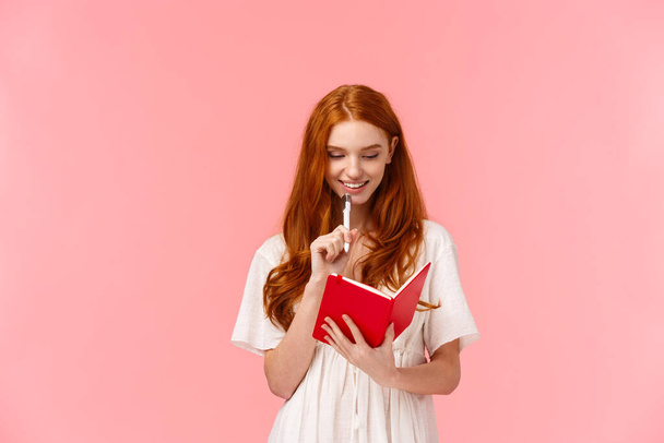 Bedachtzaam en gelukkig, glimlachend mooie roodharige vrouw in witte jurk, aanraken kin met pen tijdens het denken, het maken van kruidenier of to-do lijst, het schrijven van iets in schattige rode notitieboek, roze achtergrond - Foto, afbeelding