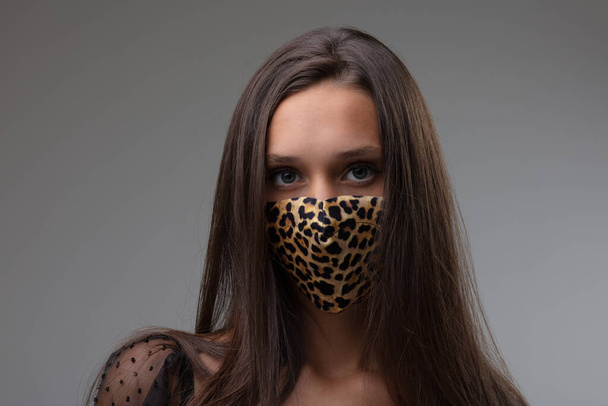 Mulher vestindo uma máscara facial de impressão animal na moda como proteção contra o coronavírus durante a pandemia de Covid-19 contra um fundo cinza em um retrato frontal
 - Foto, Imagem